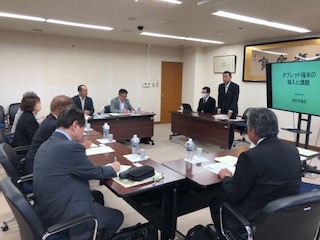 室内前方に資料が映し出された大きなモニターが設置され、コの字に設置された長机の席に議員の方々や関係者が座り、モニターの左側の席から立ち上がった男性の話を資料を見ながら熱心に聞いている視察研修の様子の写真