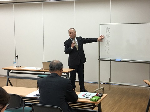 「議員憲法」と書かれたホワイトボードを指し、マイクを持って話をする田口先生の写真