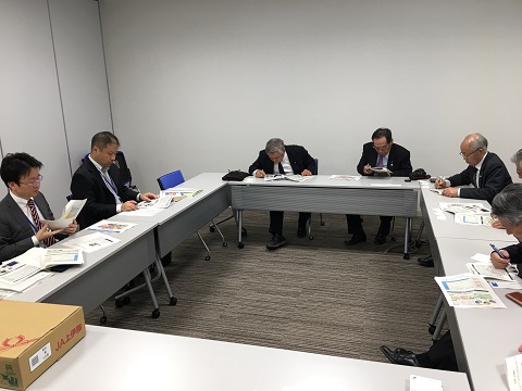 四角になるように長机を配置し資料を見ながら説明を聞く関係者の方々の写真