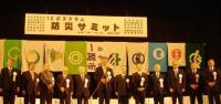 「防災サミット」と書かれた横断幕が掲げられ、箕輪町と豊島区など防災協定締結を結んだ都市の市章が並んだ舞台に立っているサミット参加者の写真