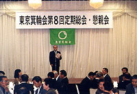 「東京箕輪会第8回定期総会・懇親会」と書かれた幕が掲げられた舞台に立っている男性と席についている参加者の写真