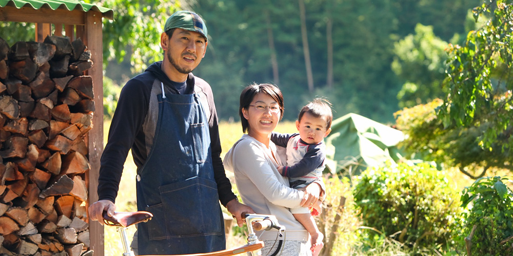 屋外の薪が積まれている場所の前で自転車と一緒に写っている安田さんと男の子を抱いている奥様の写真