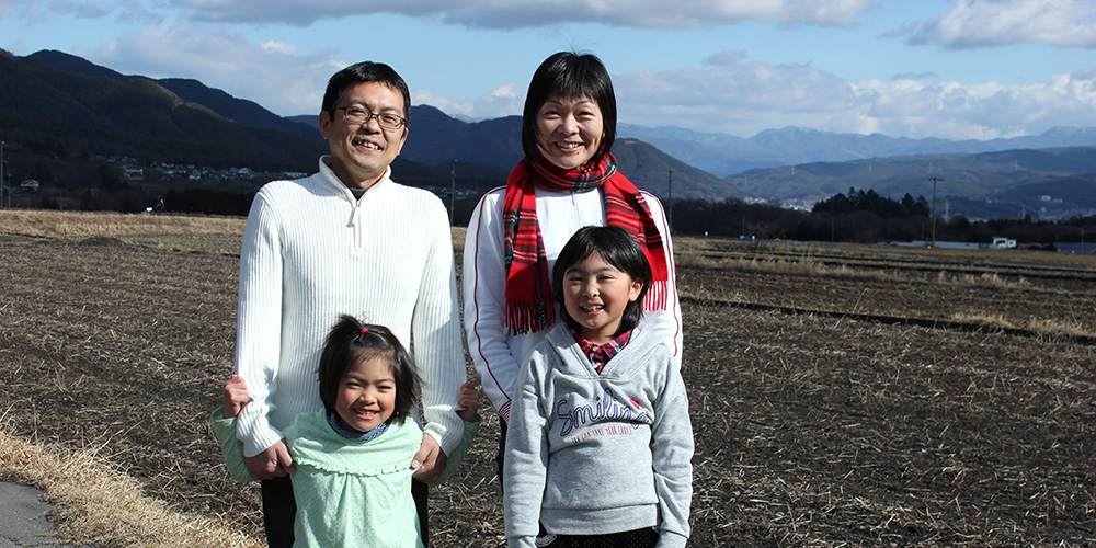 遠くに見える山々と広々とした畑の前に立っている城内さんと奥様、二人の子供の写真