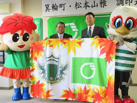 箕輪町の町章と町の観光名所「もみじ湖」の紅葉をデザインしたタイアップフラッグを持っているガンズくんともみじちゃん、その間に立っている白鳥政徳町長と神田文之社長の写真