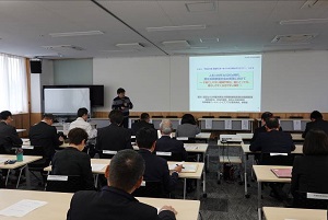 席に座り壁に映し出されている資料を見ている参加者達を室内の後方から撮影した写真