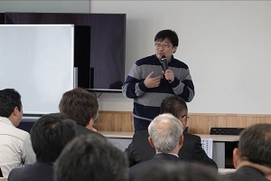 ボーダー柄の服を着た男性が参加者達の前でマイクを持って話をしている写真
