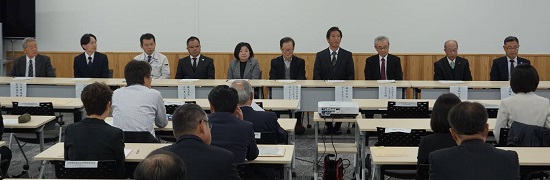 机に紙が貼られた前方の席に並んで座っている10名の関係者達の写真