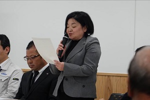 グレーのジャケットを着た女性が右手でマイクを持ち、左手に持った紙を読み上げている様子の写真