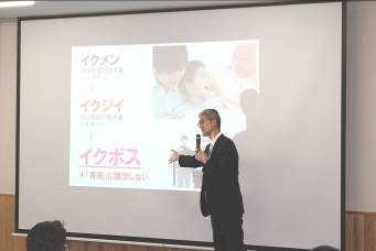 「イクメン」「イクジイ」「イクボス」の文字が映し出されたスクリーンの横で男性がマイクを持ち話をしている写真