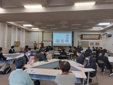 参加者達がそれぞれグループに分かれて席に着き、スクリーン左側でマイクを持って話している男性の話を聞いている様子の写真