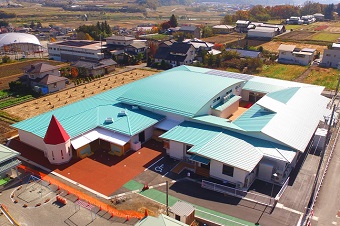 一部に円錐状の赤い屋根で、全体がエメラルドグリーン色の屋根をした沢保育園全体を上空から撮影した写真