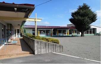 園庭の中央付近に大きな1本の木があり、園庭に沿ってL字型に園舎が建っている上古田保育園の外観写真
