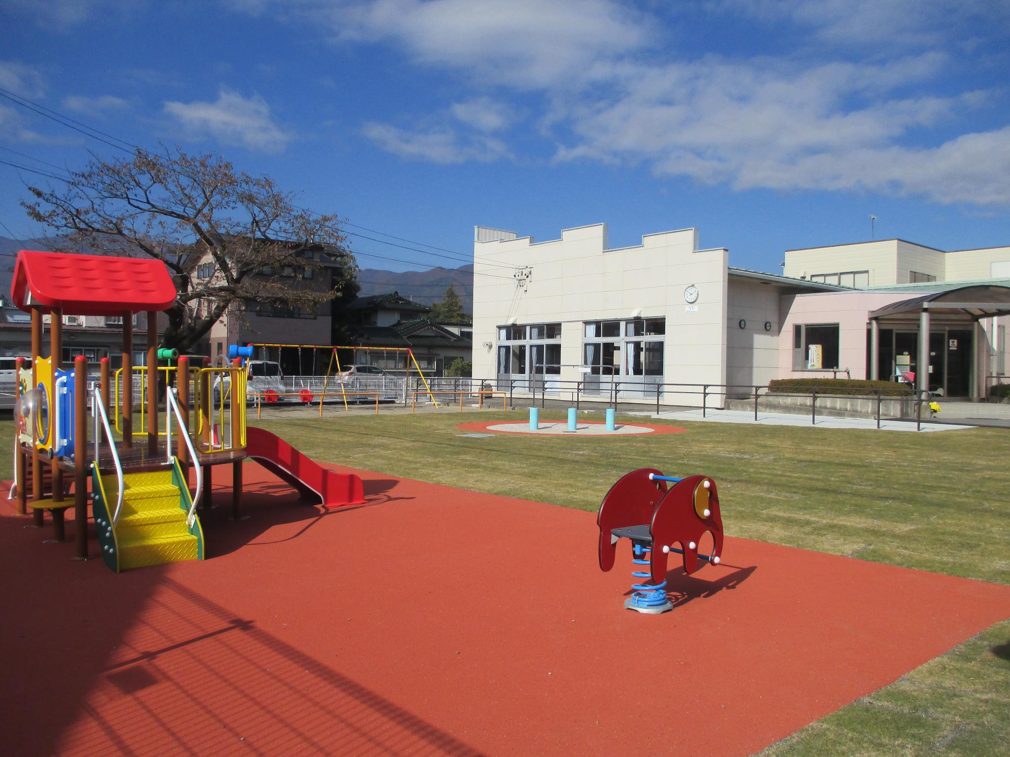 複合遊具とゾウのスプリング遊具が芝生の広場に設置され、奥に子育て支援センター「いろはぽけっと」の建物が写っている写真