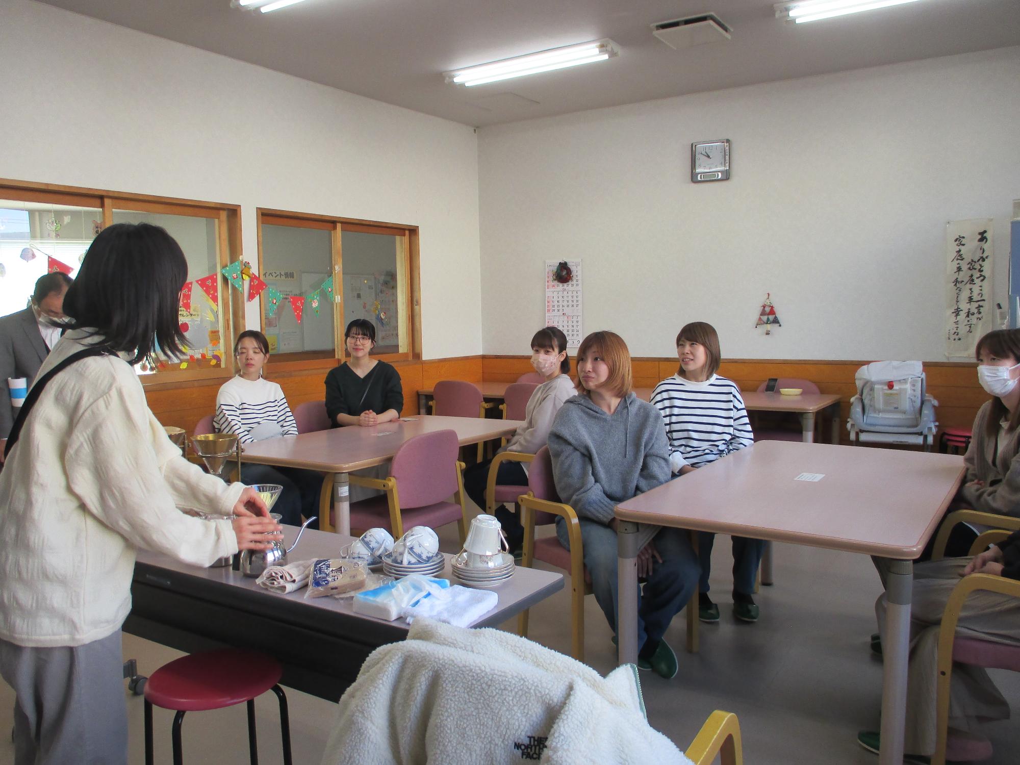 6人の椅子に座った参加者がコーヒーを入れている女性をみている