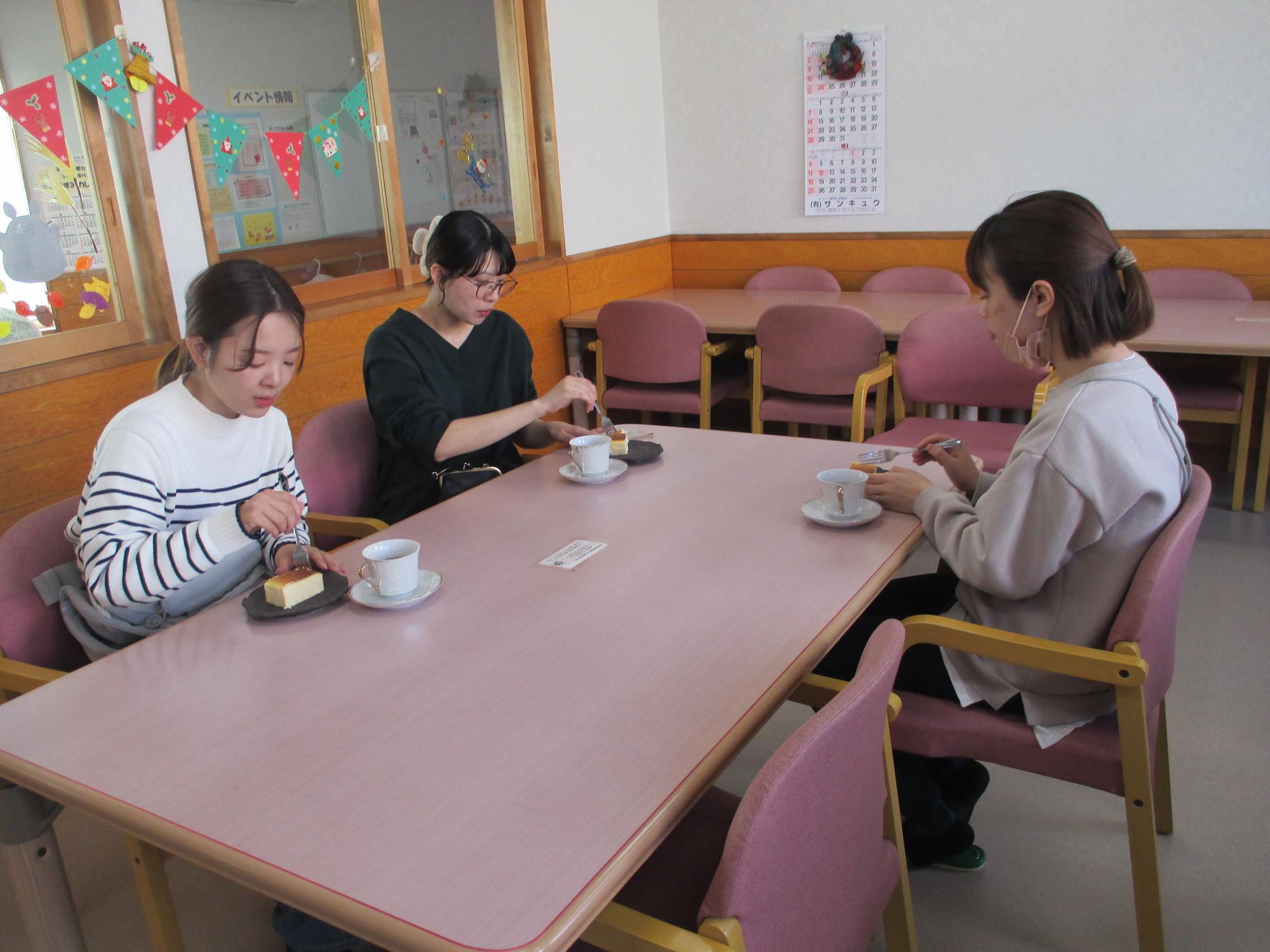 3人の女性が1台のテーブルを囲んで座り机上のチーズケーキを食べようとしている。チーズケーキの隣にコーヒーカップが置いてある。
