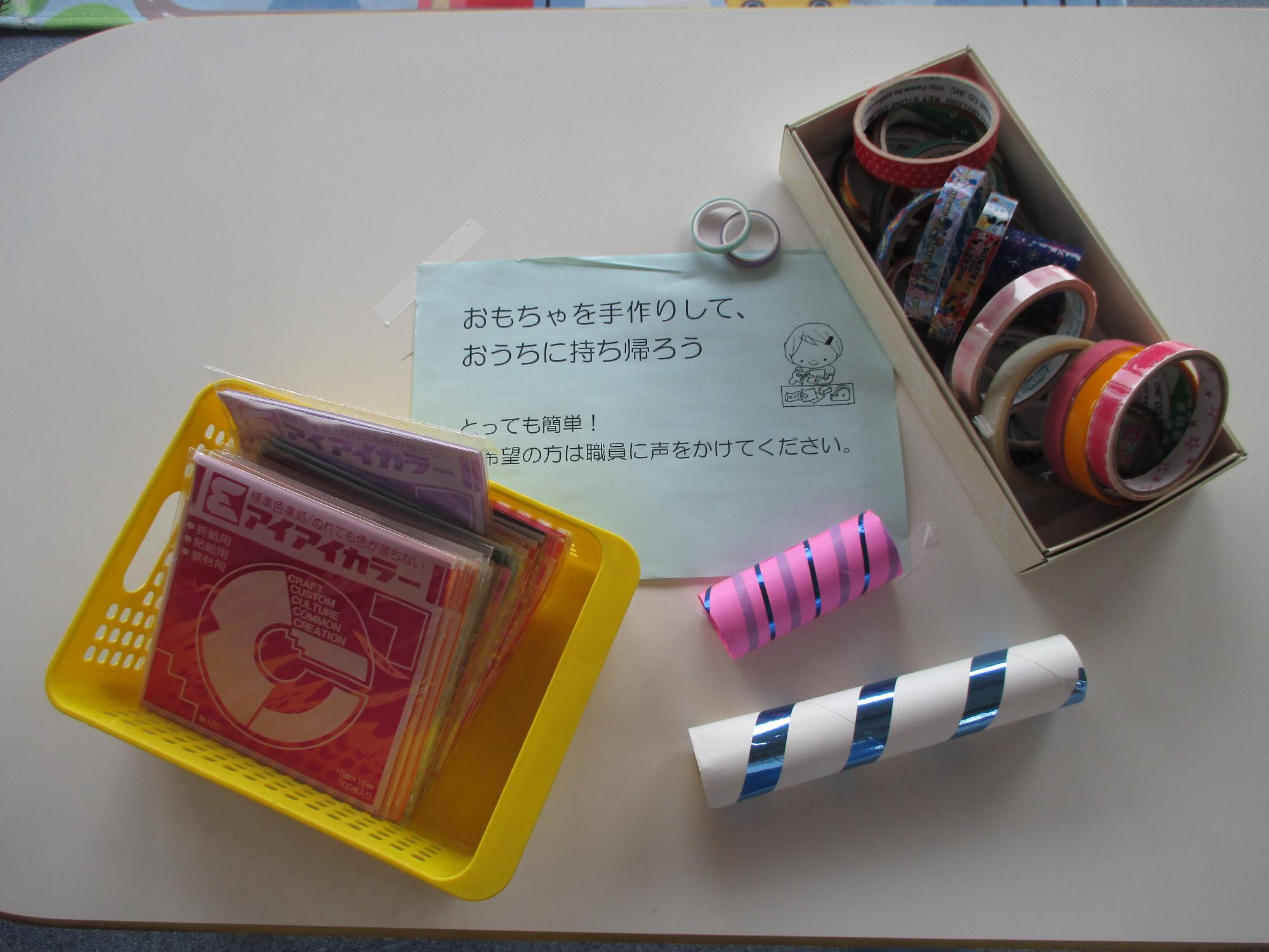 色紙、カラフルなマスキングテープ、デコレーションしたペーパータオルの芯とおもちゃを手作りしておうちに持ち帰ろうの張り紙