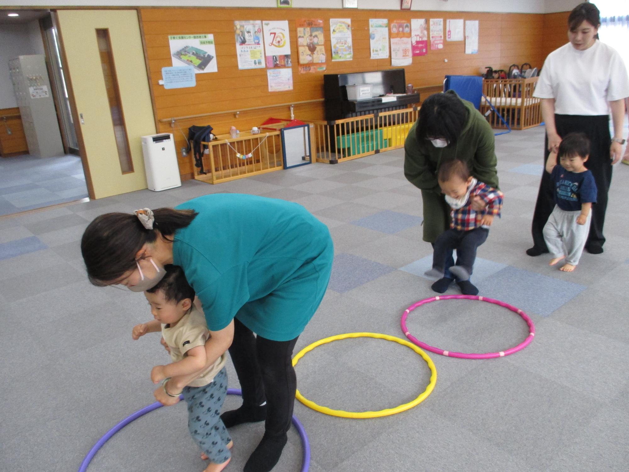 1列に並んだフープのところを両足ジャンプがまだできない子が母に抱えられてジャンプしている