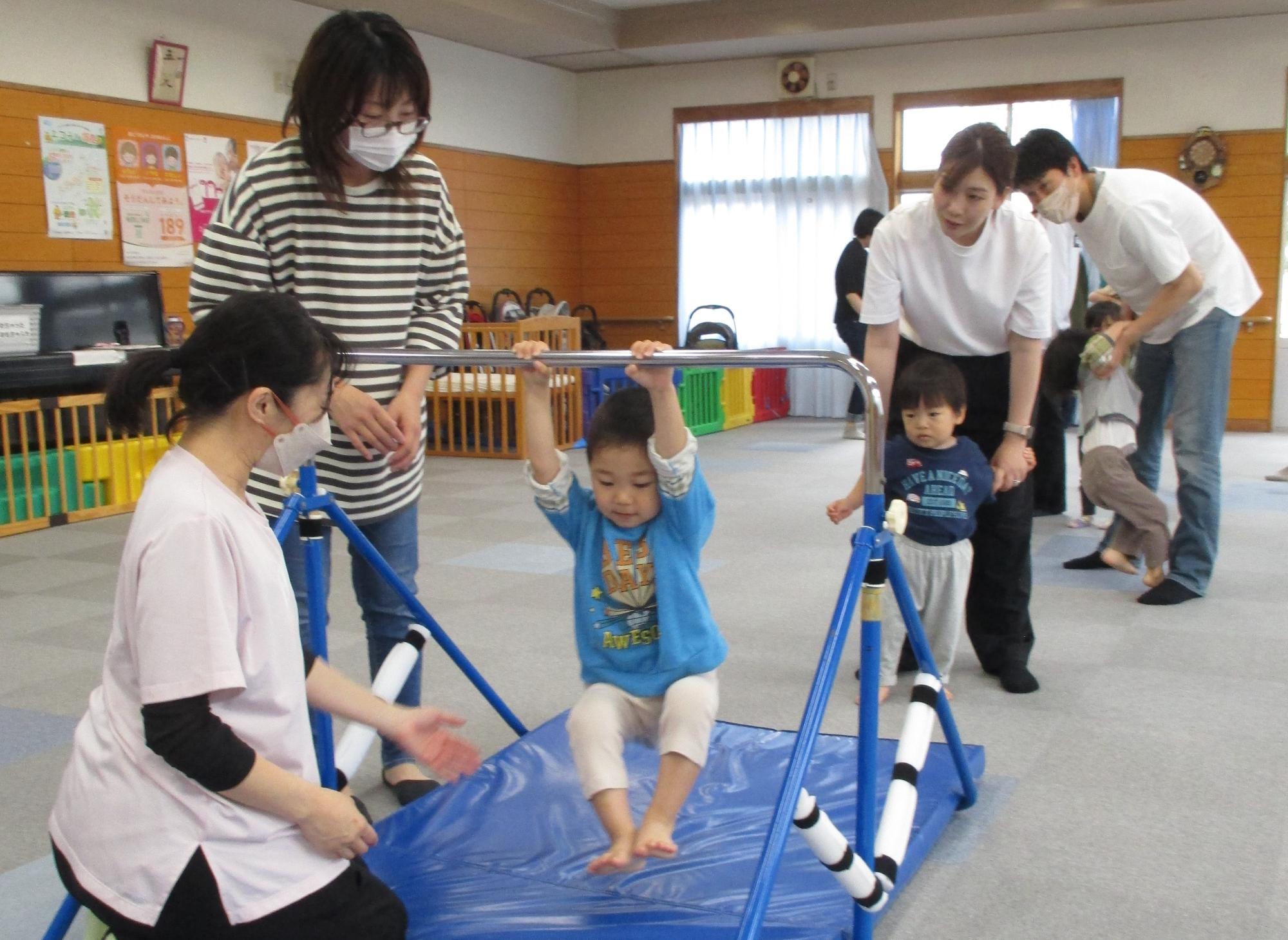 鉄棒に両手でぶら下がっている男の子横に母が立ち、講師が膝をつき補助体制