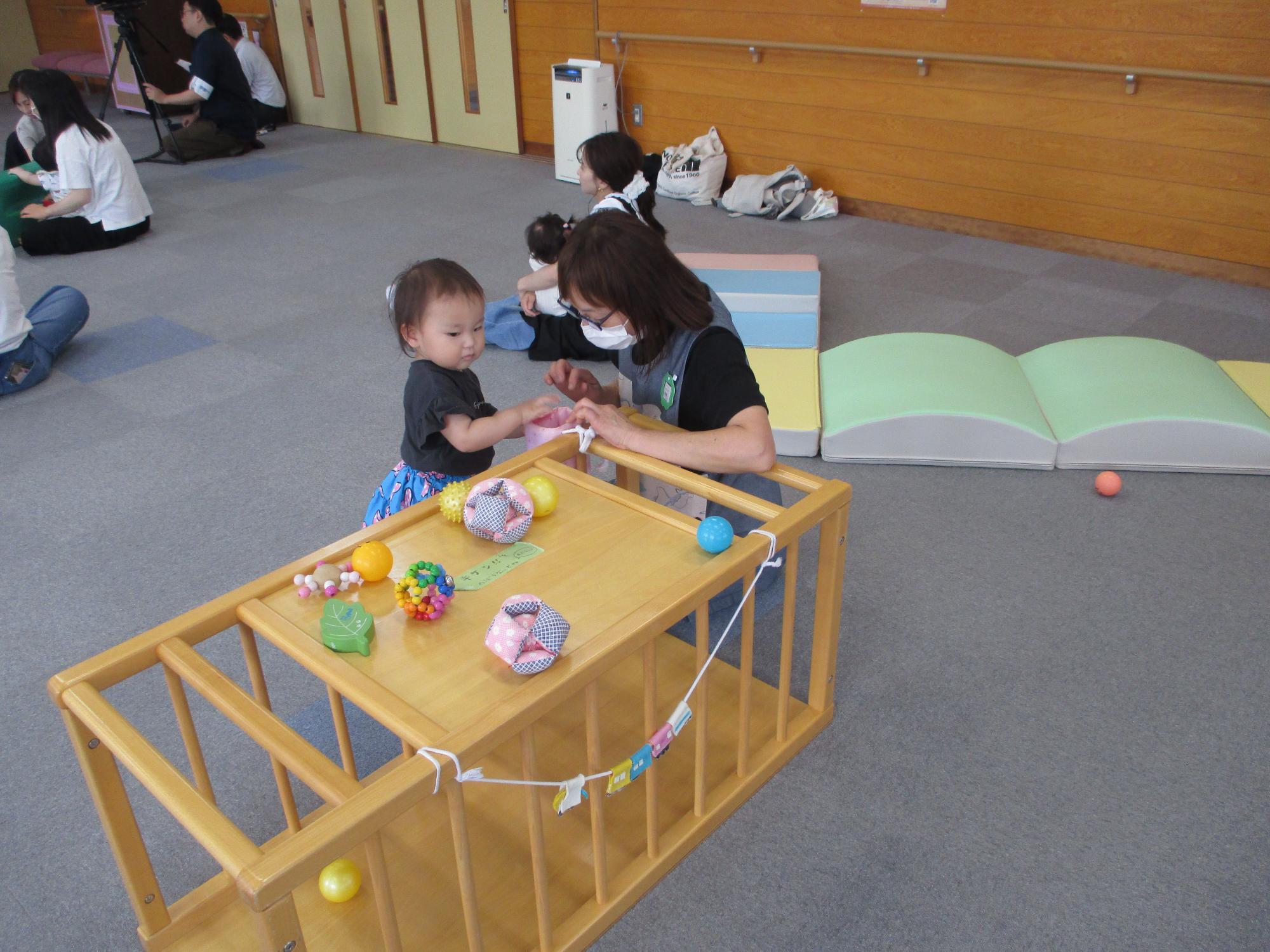 木製トンネルの上にボールがのっているところで遊んでいる女児とそれを見守るサポーター。