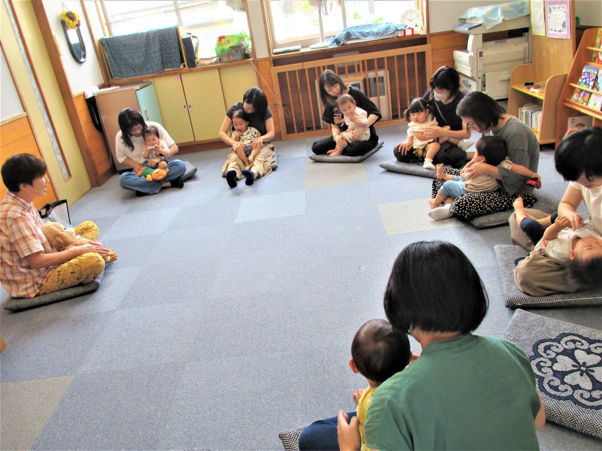 1歳くらいの子どもを膝に乗せた母子7組が講師を囲んでくるまざに座っている