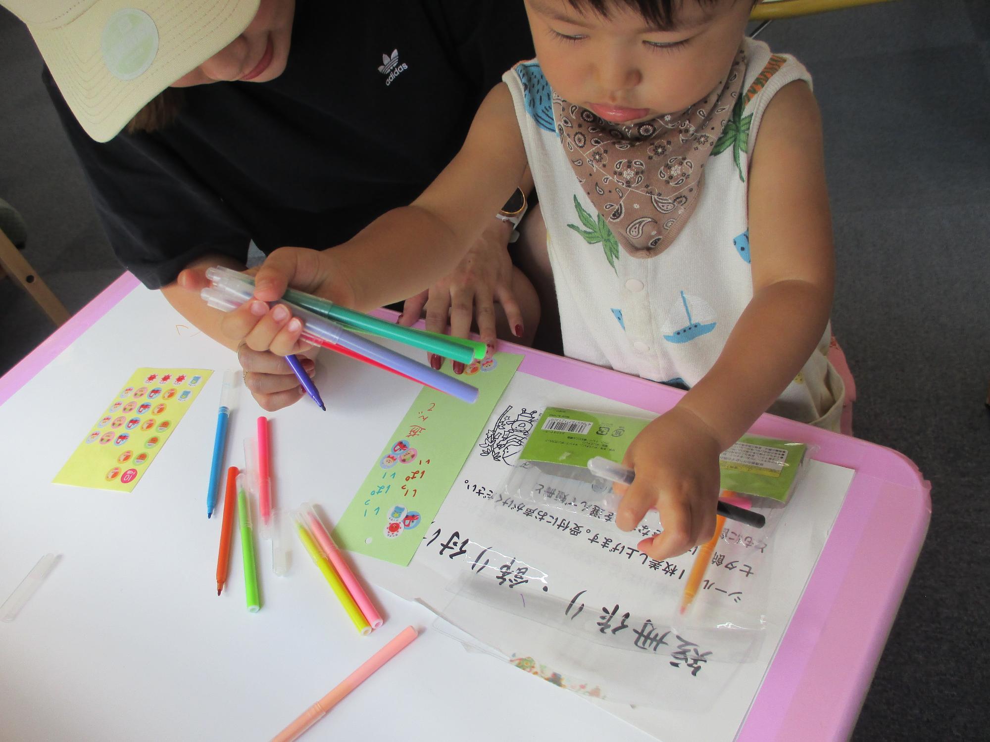 短冊に願い事を書いている母親と右手に4本のカラーペンを握り占めてみている1才男児
