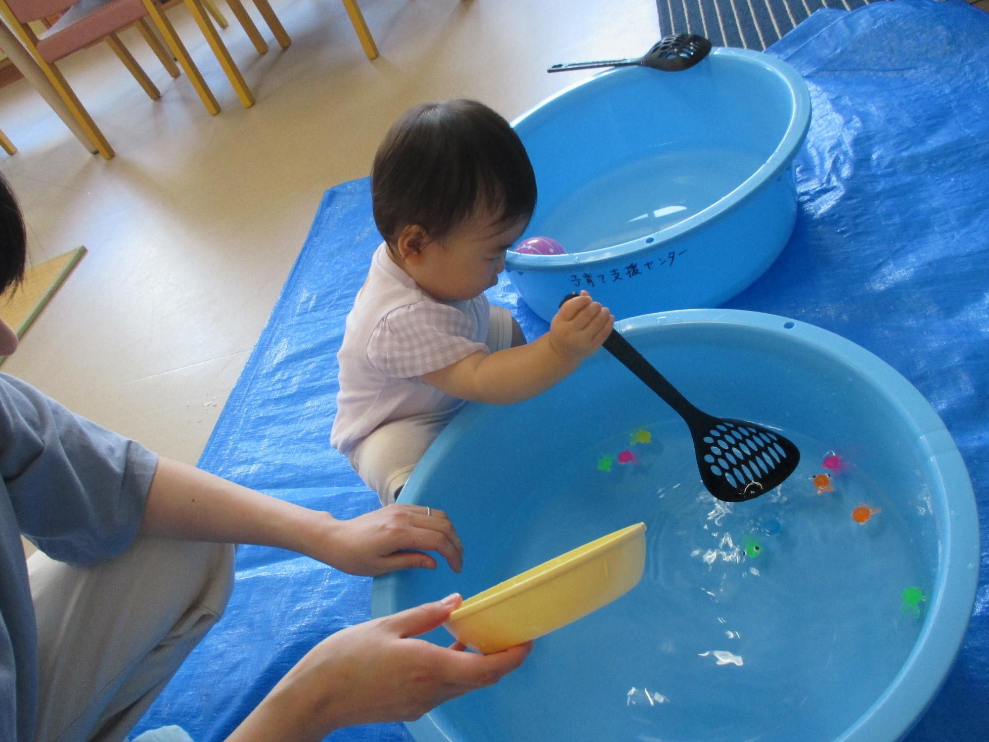 水を張ったタライに浮かぶおもちゃの金魚を穴あきオタマですくおうとしている1歳女児