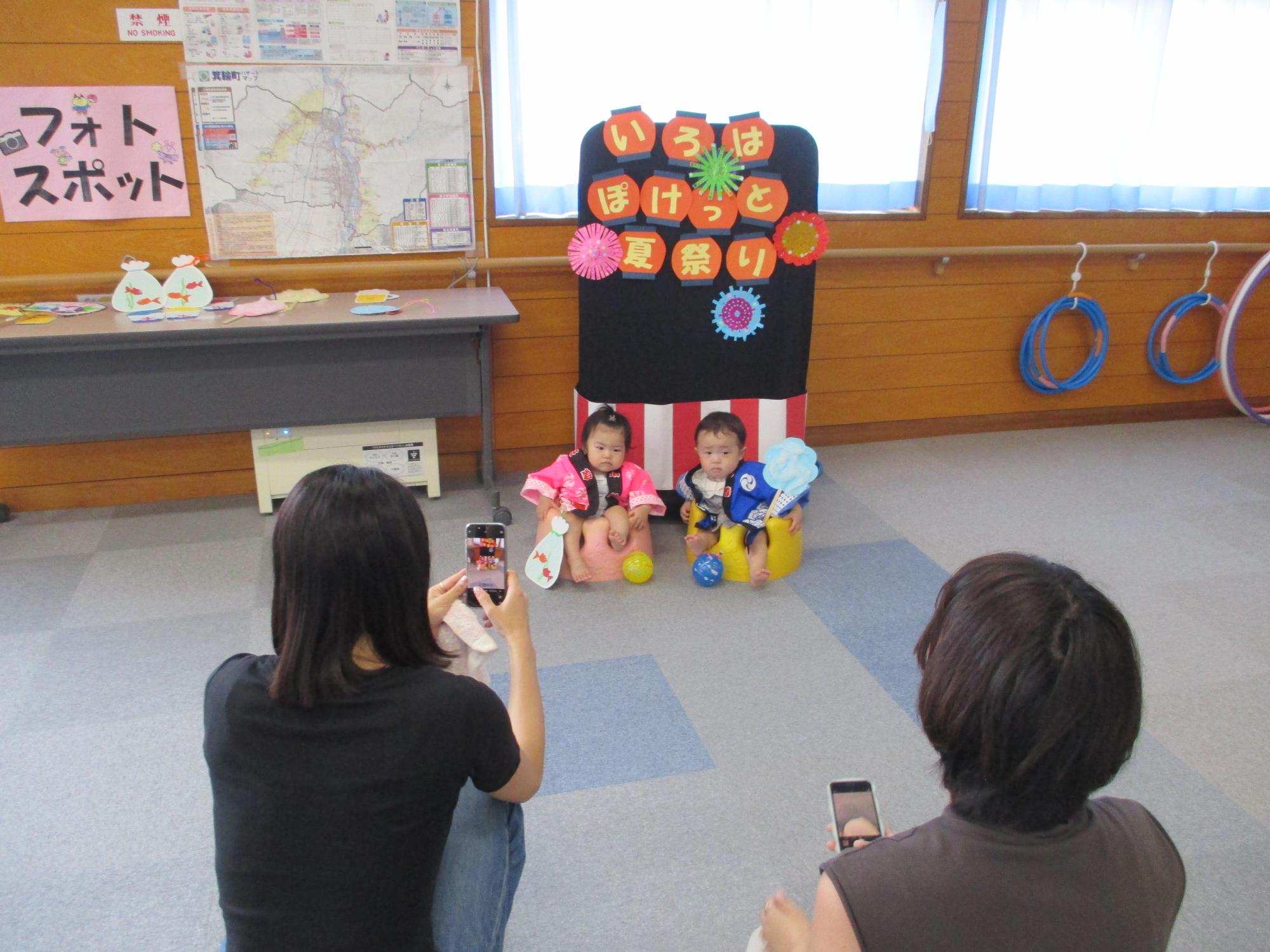 「いろはぽけっと夏祭り」と書いてあるフォトスポットの前で祭り法被姿で並んで座る2人の1歳児をスマホで撮影している母
