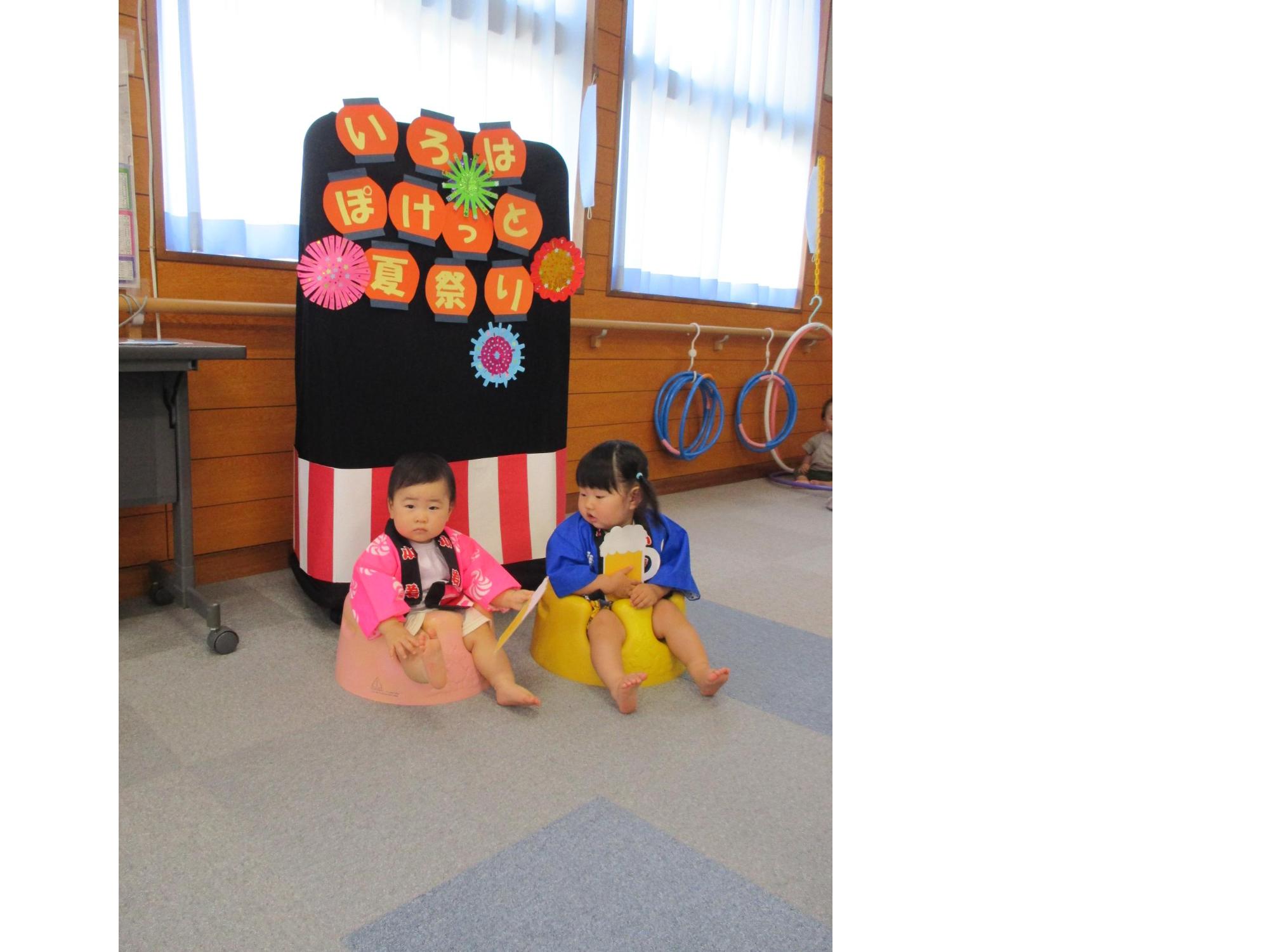 「いろはぽけっと夏祭り」と書いてあるフォトスポットの前で祭り法被姿で並んで座る2人の1歳児