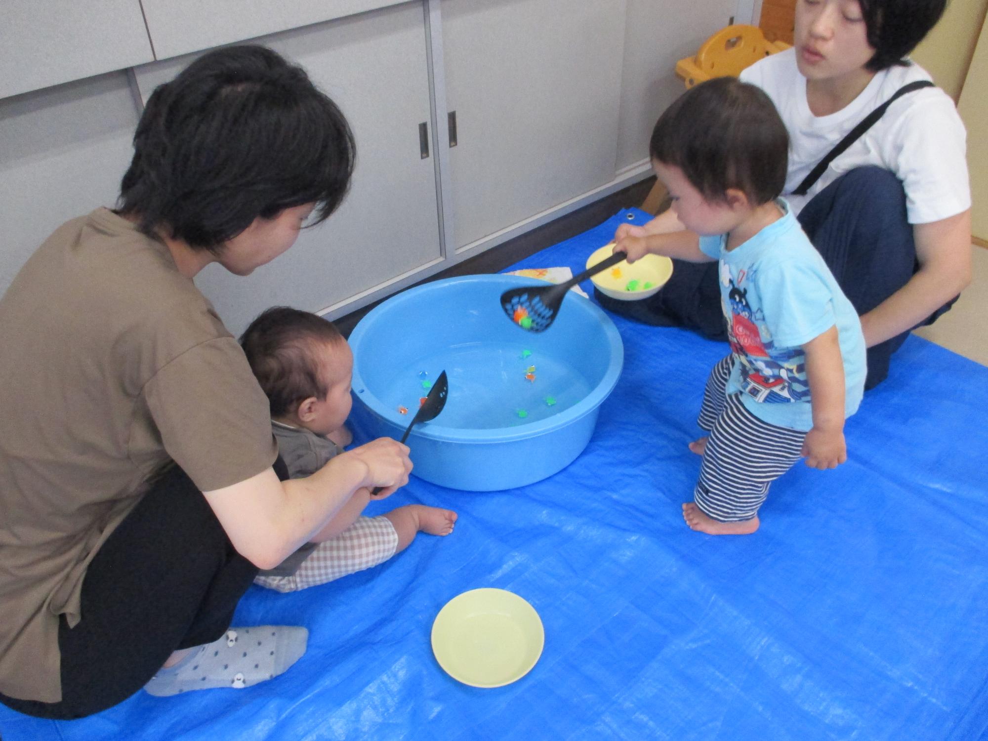 水を張ったタライに浮かぶおもちゃの金魚を穴あきお玉ですくおうとしている1歳男児を見ている母と親子1組