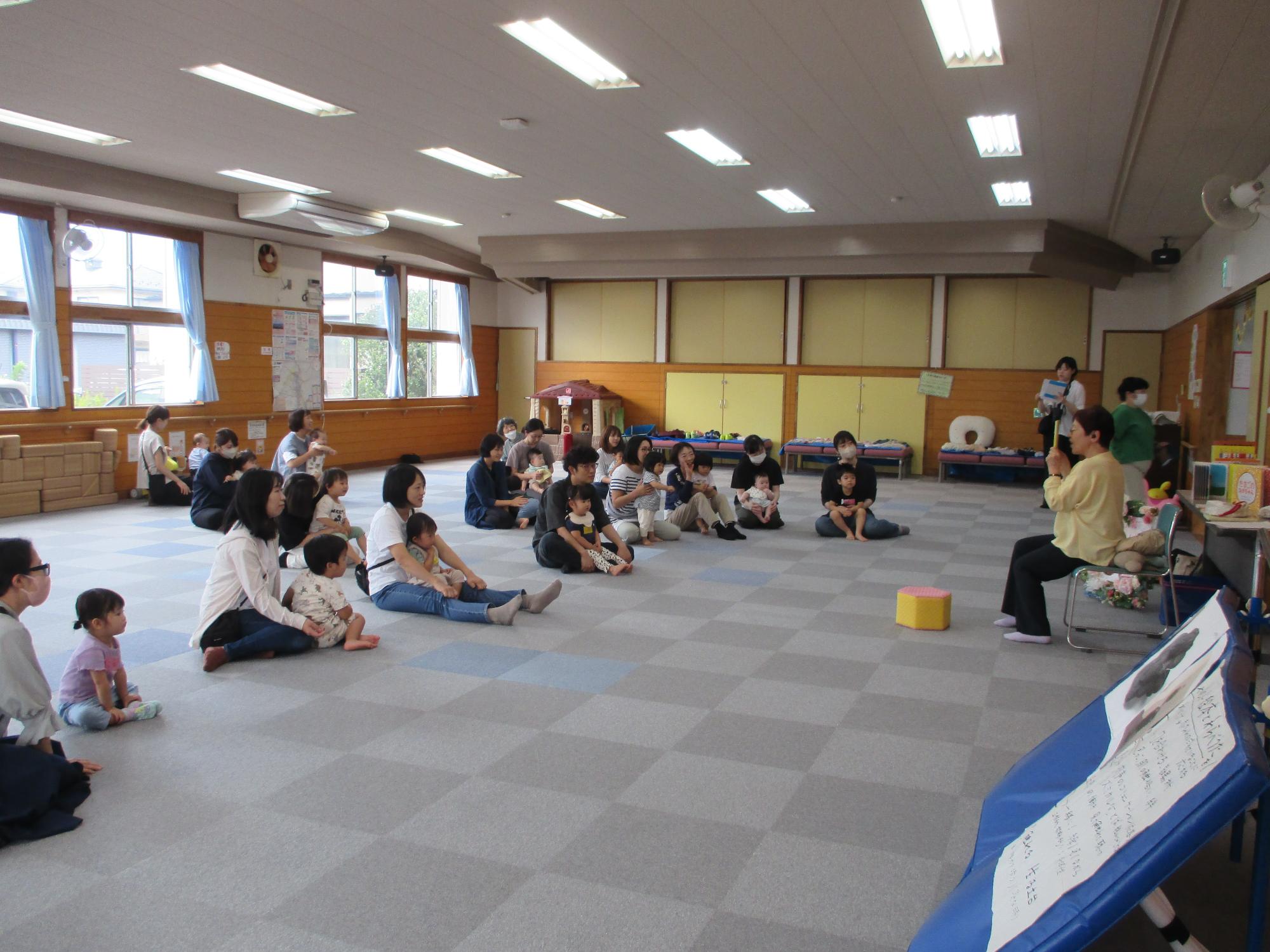 講師が絵本の読み聞かせ、大勢の親子が床に座り見聞きしている。