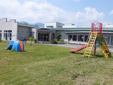 滑り台や太鼓梯子が芝生の広場に設置され、奥に子育て支援センター「いろはぽけっと」の建物が写っている写真
