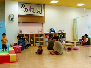 マットが敷かれた場所で遊んでいる男の子、床をハイハイしている子供を見守っているお母さん、一緒に絵本を読んでいる様子の親子が子育て支援センター「みのわ～れ」ほっこりルームを活用している写真