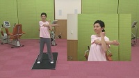黒色のマットの上に立つインストラクターの女性が足を肩幅に開き、腕を体の前で交差し左腕を体に引き付けている全体と上半身の映像の写真（立って行うストレッチのサイトへリンク）