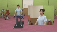 黒色のマットの上に立つインストラクターの女性が足を肩幅に開き、青色のタオルを両手で持っている全体と上半身の映像の写真（立って行うストレッチ（ソフトタイプ）のサイトへリンク）