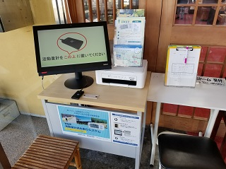正方形の台の上にパンフレット、プリンターとモニターが設置され、画面に活動量計の写真と「活動量計をこの上に置いてください。」と書かれた文字が表示されている写真