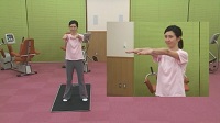 黒色のマットの上に立つインストラクターの女性が足を肩幅に開き、腕を前に揃えて伸ばしている全体と上半身の映像の写真（立って行う体操のサイトへリンク）