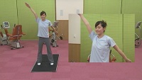 黒色のマットの上に立つインストラクターの女性が足を肩幅に開き、右手を斜め上に上げ左手を斜め下に下げている全体と上半身の映像の写真（立って行う体操（ソフトタイプ）のサイトへリンク）