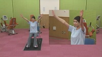 黒色のマットの上に設置された椅子に座ったインストラクターの女性が足を肩幅に開き、両手を上に上げ万歳している全体と上半身の映像の写真（座って行う体操のサイトへリンク）