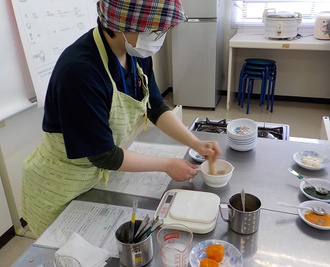 チェック柄の三角巾を頭に巻き、黄緑色のエプロンを着用した女性が、取っ手が付いた白色の器にすりこぎでゴマをすり離乳食を作っている様子の写真