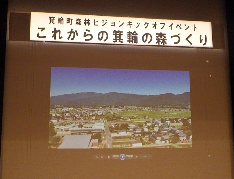 スクリーンに「箕輪町森林ビジョンキックオフイベント これからの箕輪の森づくり」と書かれ、山を背景にした街並みが映し出されている写真