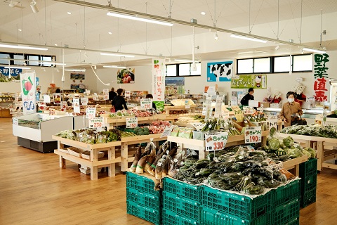 広い室内にコンテナや棚に多くの野菜が並べられお客さんが買い物をしている写真