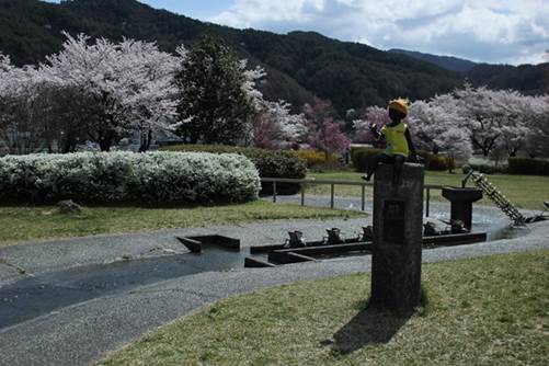 桜とつつじの花が満開に咲き小川が流れ、子供の石像がある公園の写真