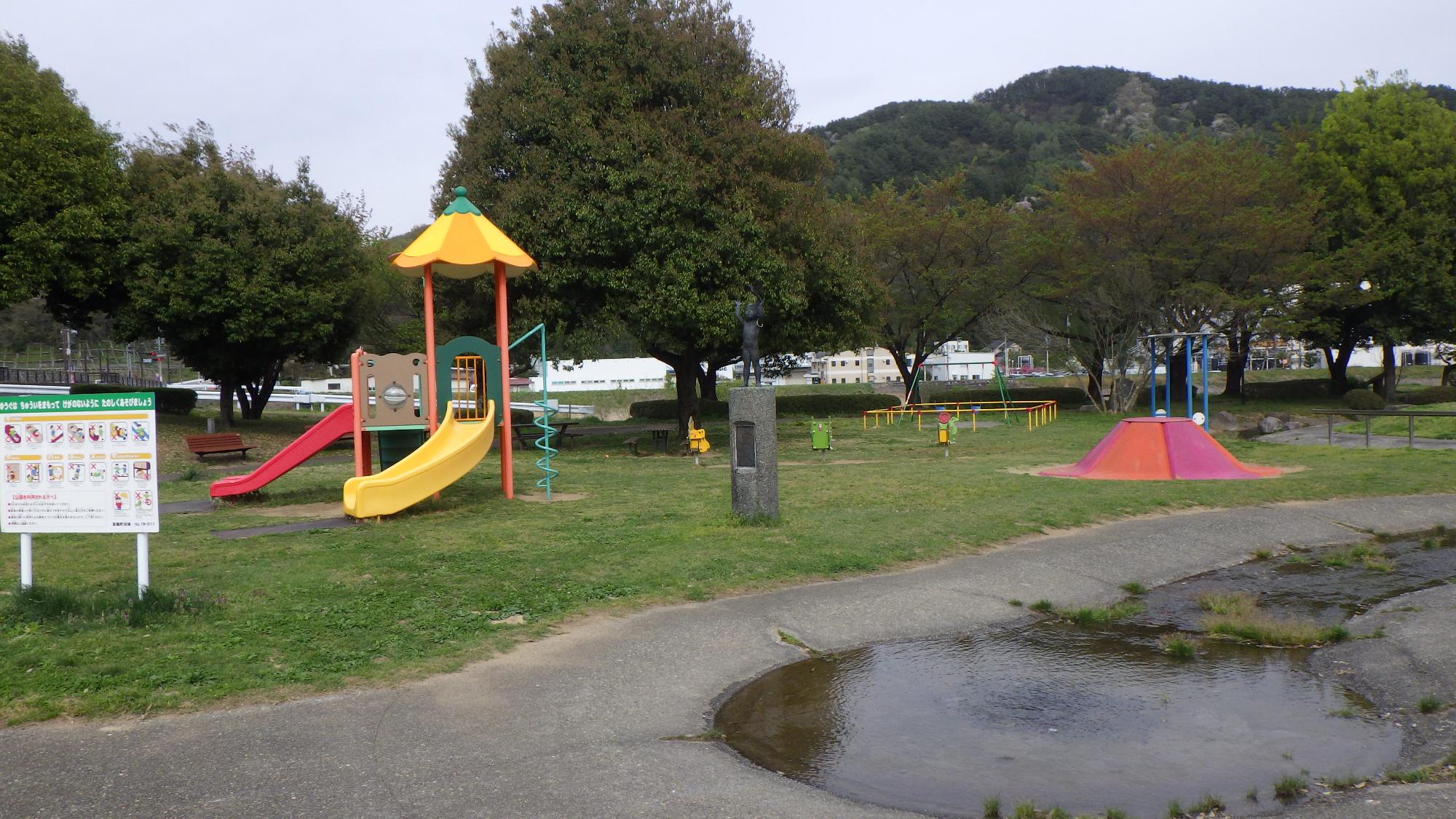 木々と山をバックに滑り台やブランコなどの遊具が芝生の上にあり、手前に水辺がある写真