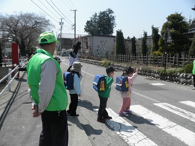 横断歩道を渡っている子どもたちを緑色のジャケットを着た見守り隊の方が見守っている写真