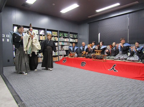 壁に本棚が設置されている館内で、右側に並んで座っている演奏者の太鼓や笛などの音色に合わせて、左側で着物を着た古田人形芝居保存会の3名の方が「三番叟」の奉納を行っている様子の写真