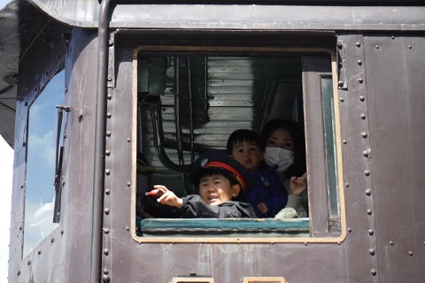 小さな子供を抱っこした母親と車掌さんの帽子を被り、運転席に座っている男の子が笑顔で汽笛体験を行っている様子の写真