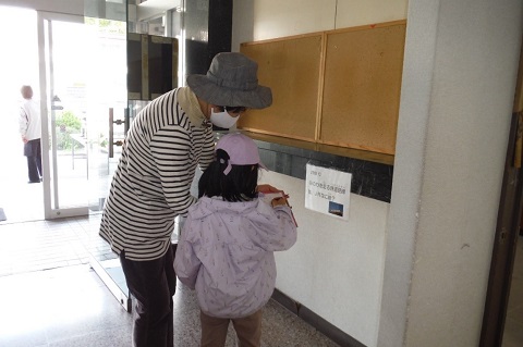 薄ピンク色のキャップにジャンパーを着た女の子とグレー色の帽子を被った女性が、建物入り口の壁に貼られた鉄道クイズを見ている写真