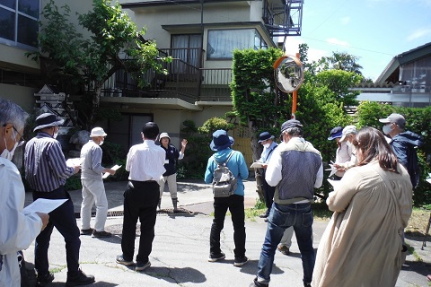 周りに人家がありカーブミラーが設置された場所に立つ職員の話を、参加者が聞いている様子の写真