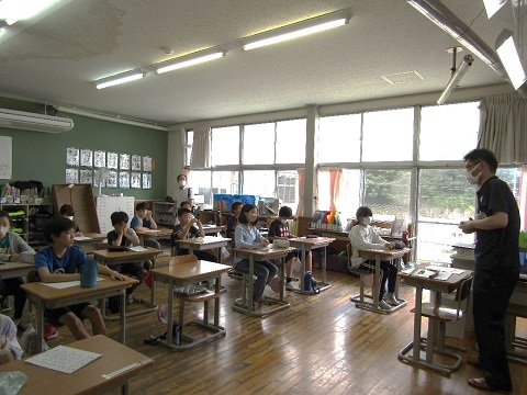 教室の前方に立つ男性職員の話を、先生や自席に座った児童たちが熱心に聞いている写真