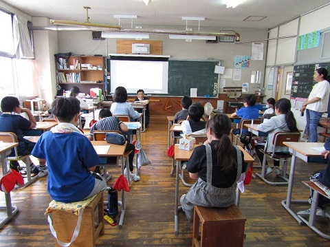 前方に設置されたスクリーンの前に座った男性職員の話を、自席に座った児童たちが聞いている後ろ姿の写真
